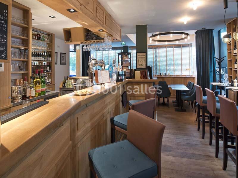 Intérieur d'un bar moderne avec comptoir en bois, tabourets, étagères de bouteilles et tables dans un espace lumineux.