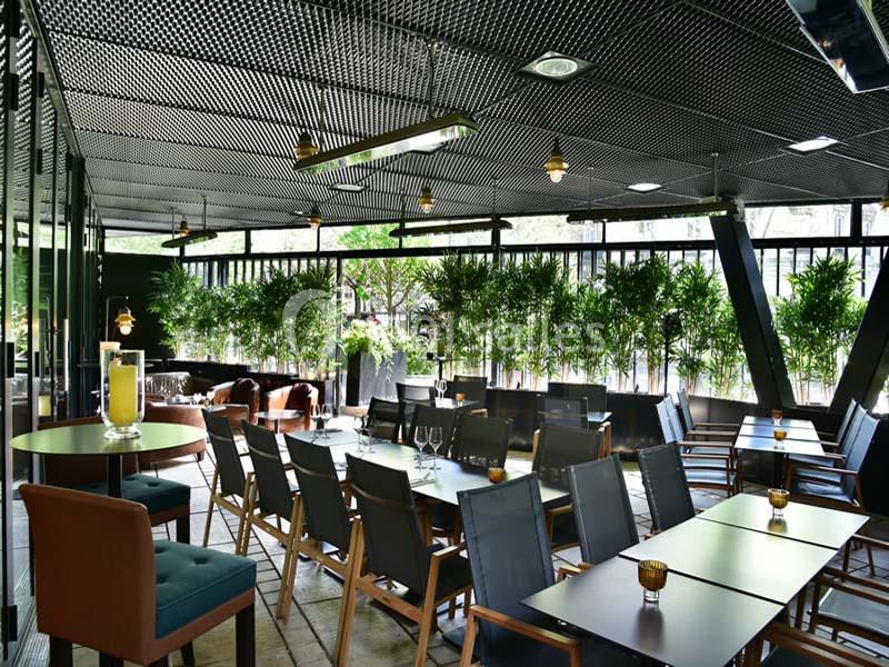 Salle de restaurant moderne avec mobilier en bois et métal, grandes baies vitrées et plantes vertes en arrière-plan.