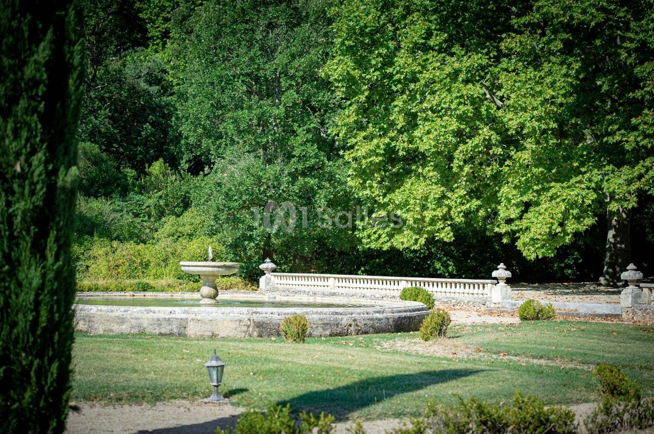 Fontaine en pierre entourée de pelouse et de buissons, avec des arbres verdoyants en arrière-plan.