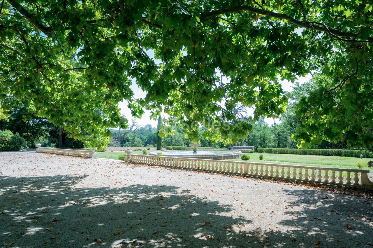 Allée ombragée par des arbres feuillus, bordée d'une balustrade blanche, avec un jardin verdoyant en arrière-plan.