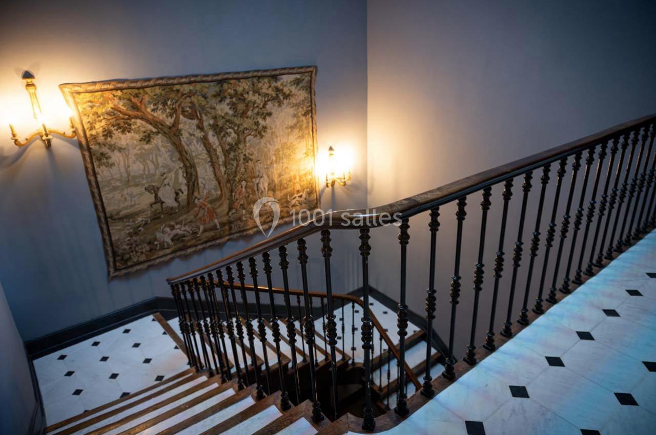 Escalier en bois avec une rampe en fer forgé, tapisseries murales et éclairage mural dans un intérieur élégant.