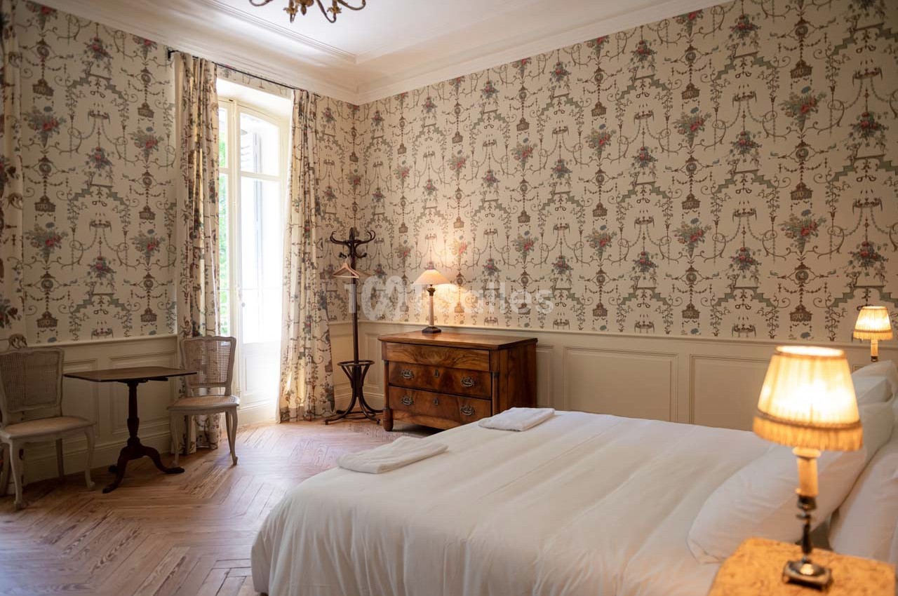 Chambre lumineuse avec lit double, commode en bois, table ronde, chaises en osier et murs ornés de papier peint floral.