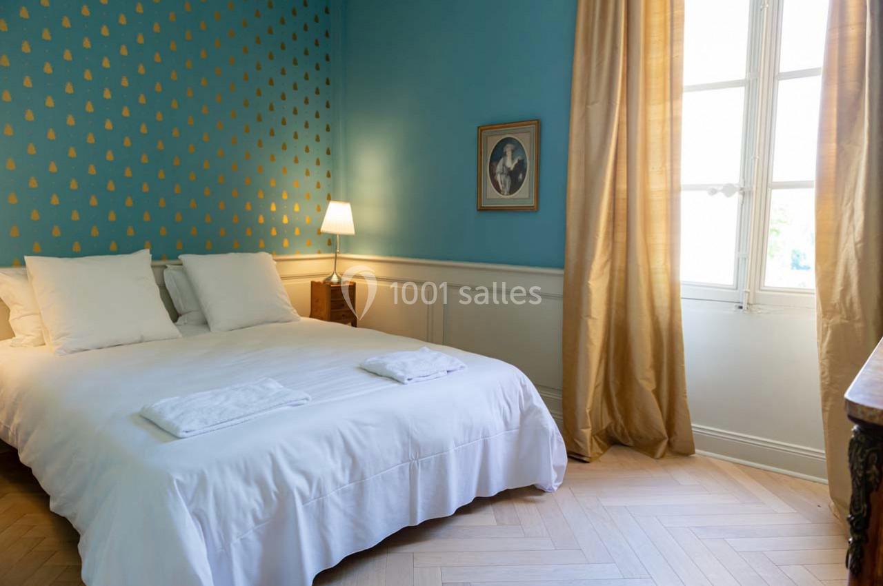 Chambre lumineuse avec un lit double, mur bleu décoré de motifs dorés, rideaux beige et fenêtre laissant entrer la lumière.