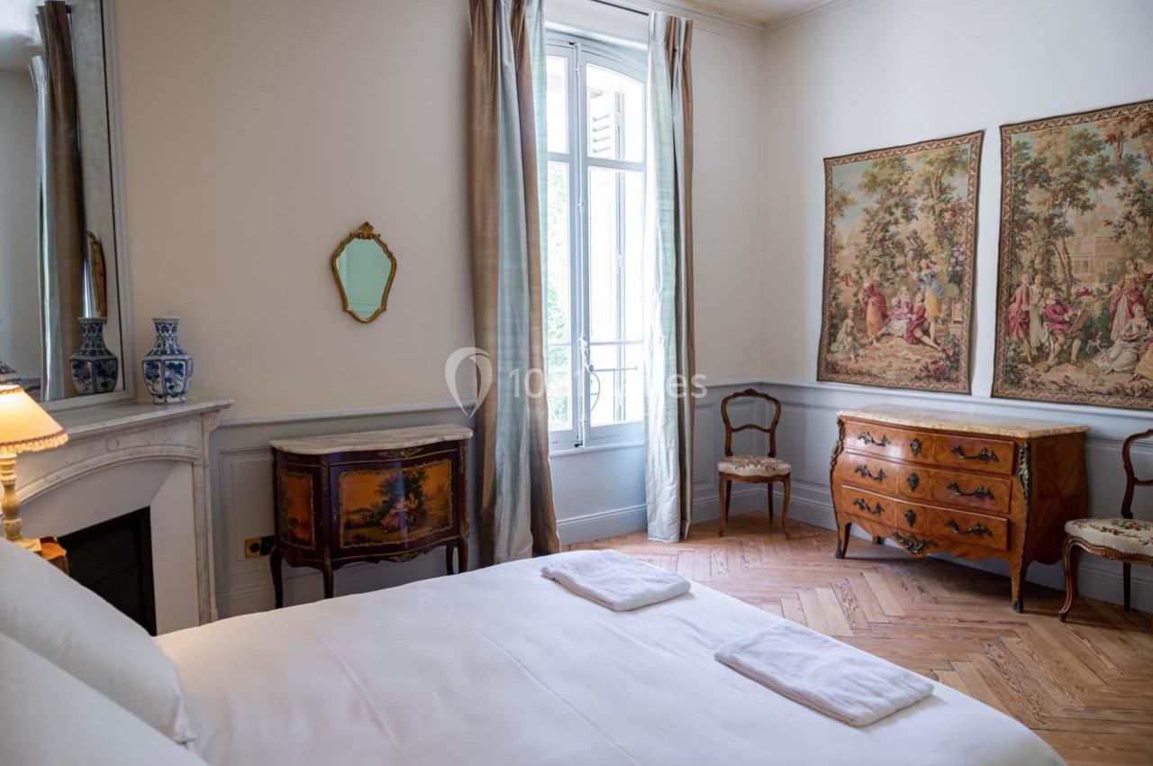Chambre lumineuse avec lit double, cheminée, meubles anciens, tapisseries murales et grande fenêtre donnant sur l'extérieur.