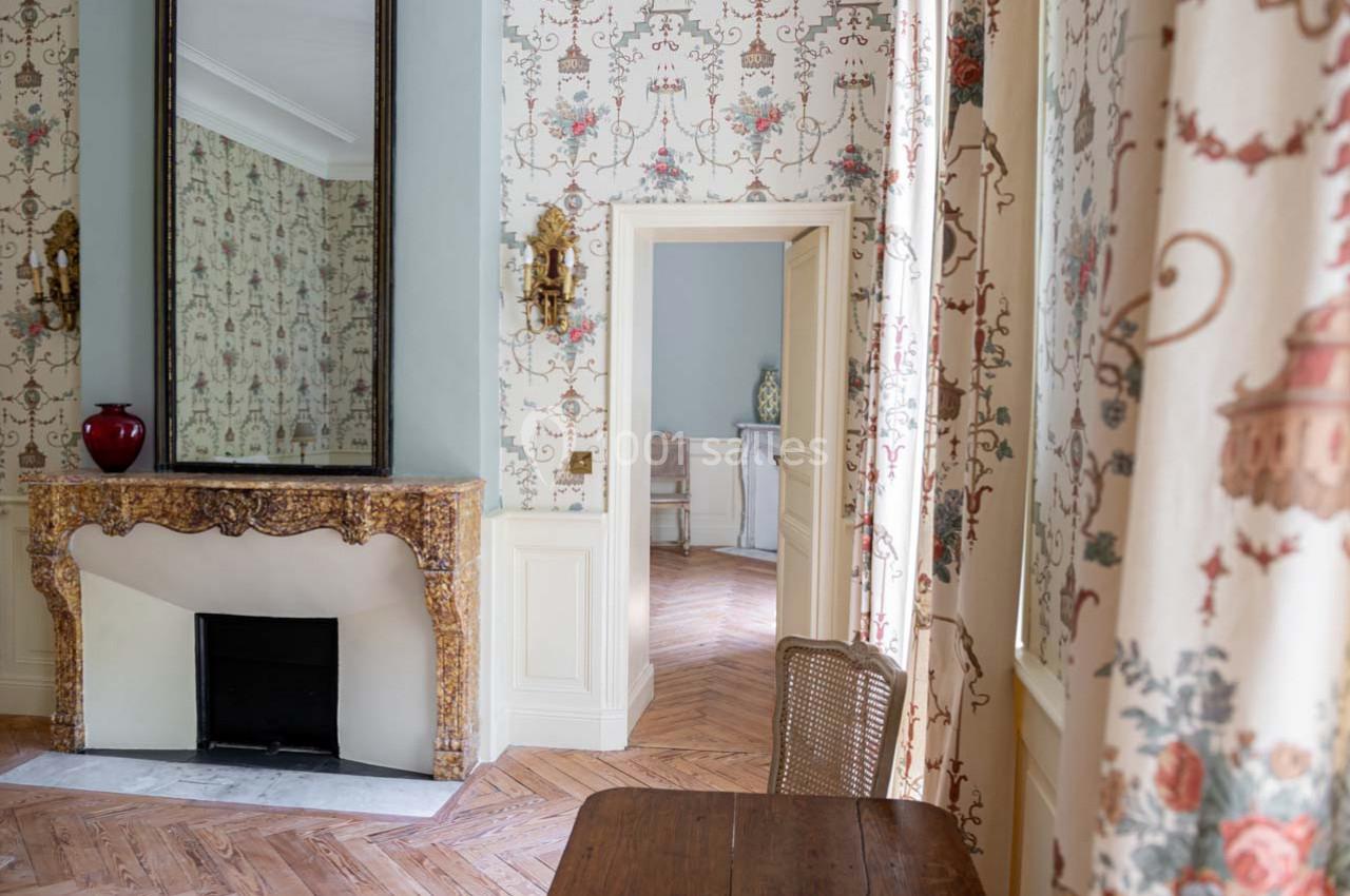 Intérieur élégant avec cheminée ornée, miroir, parquet en bois, murs tapissés et rideaux à motifs floraux.