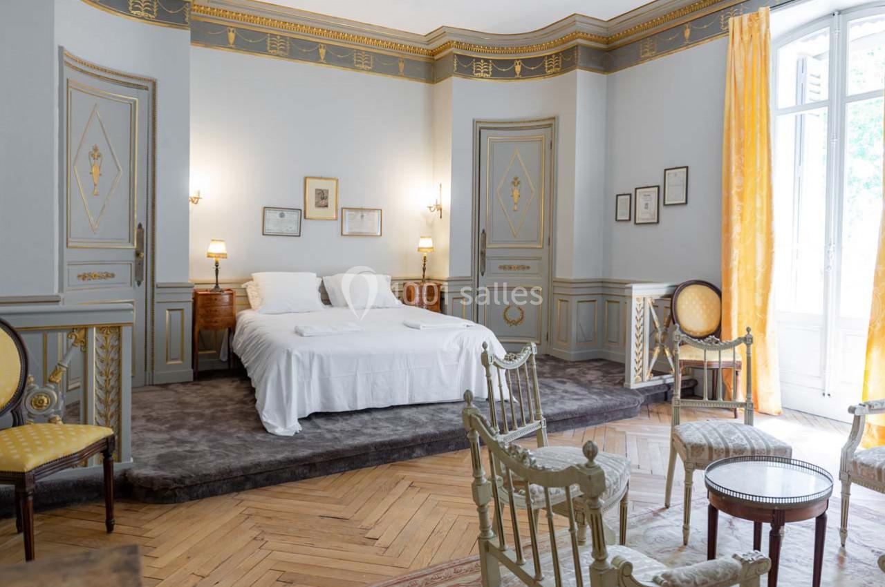 Chambre élégante avec lit double, mobilier ancien, murs ornés et rideaux jaunes, éclairée par une grande fenêtre.