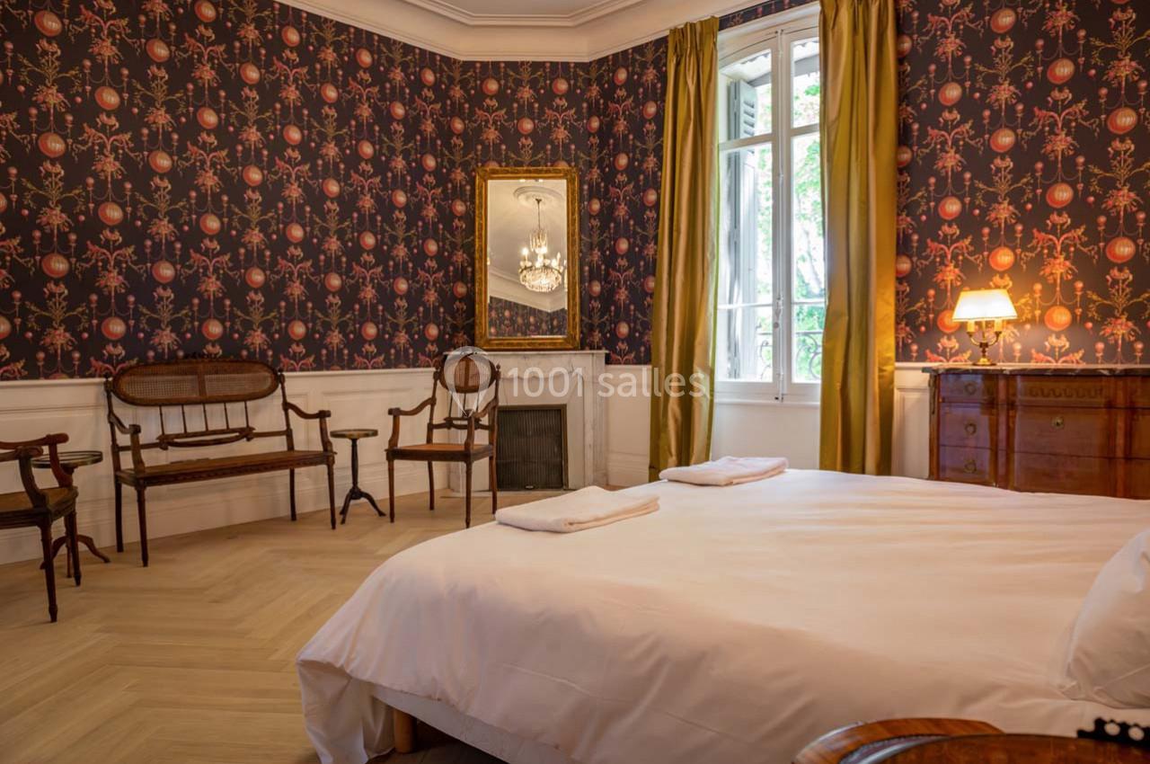 Chambre avec lit double, mobilier en bois ancien, cheminée, miroir, et fenêtre avec rideaux jaunes.