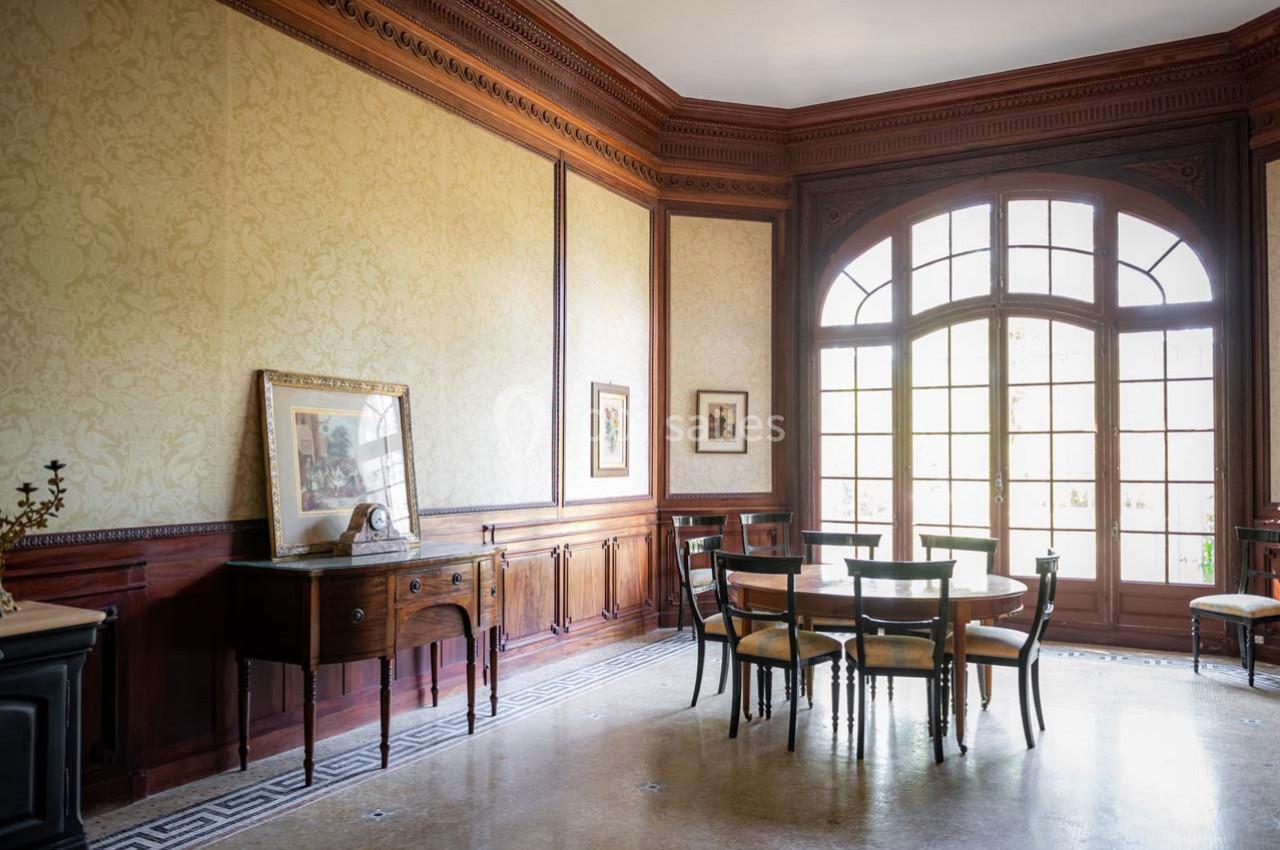 Salle à manger lumineuse avec table, chaises en bois, buffet ancien, grandes fenêtres et murs ornés de boiseries.