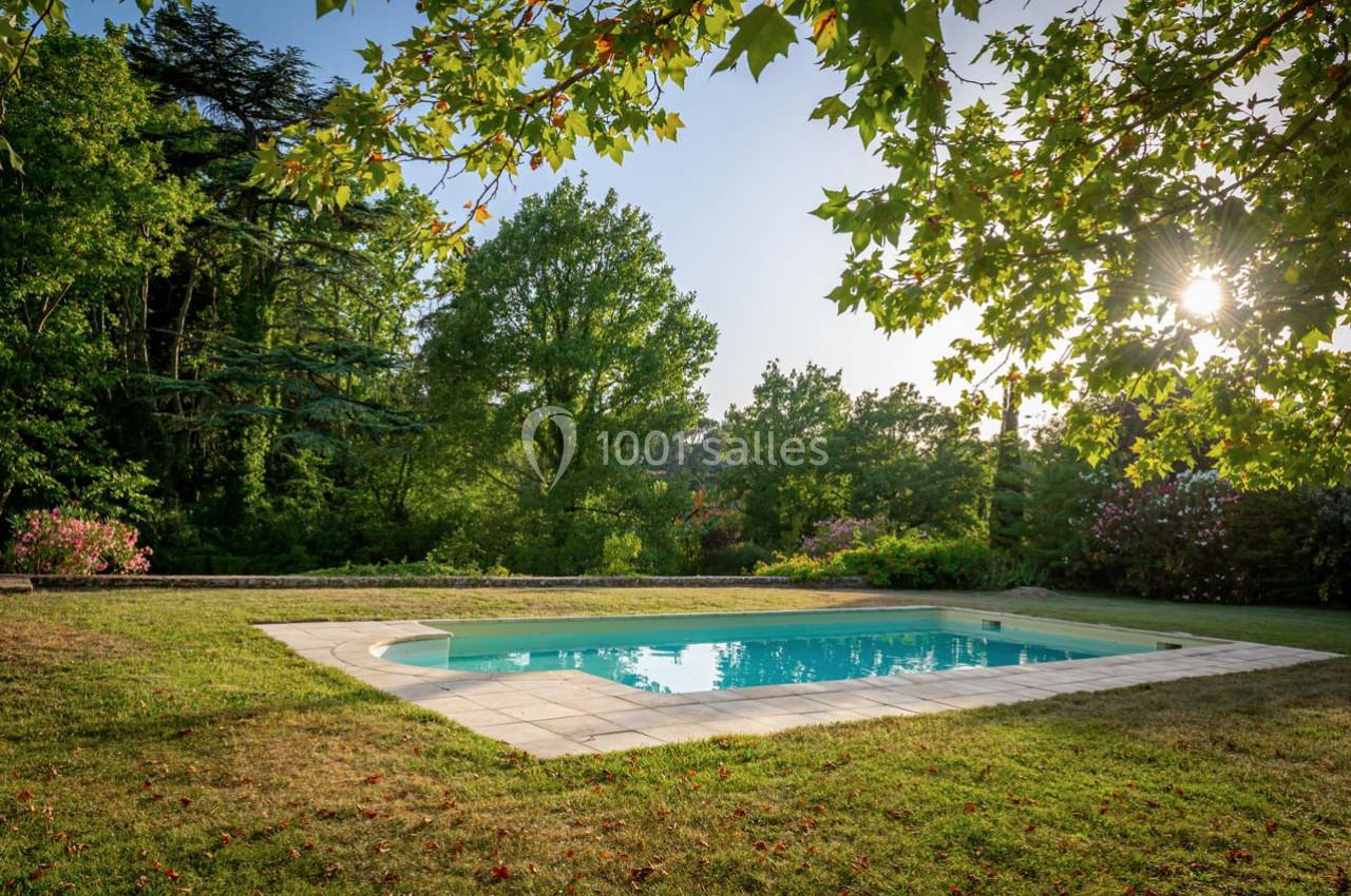 Piscine extérieure entourée de pelouse et d'arbres, sous un ciel dégagé avec le soleil filtrant à travers les branches.