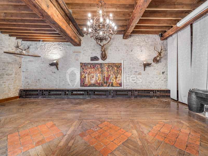 Salle avec parquet en bois, murs en pierre, tapisseries, lustre et décorations murales comprenant des têtes d'animaux.