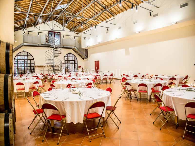 Salle lumineuse avec tables rondes dressées, chaises rouges pliantes et plafond en poutres apparentes.
