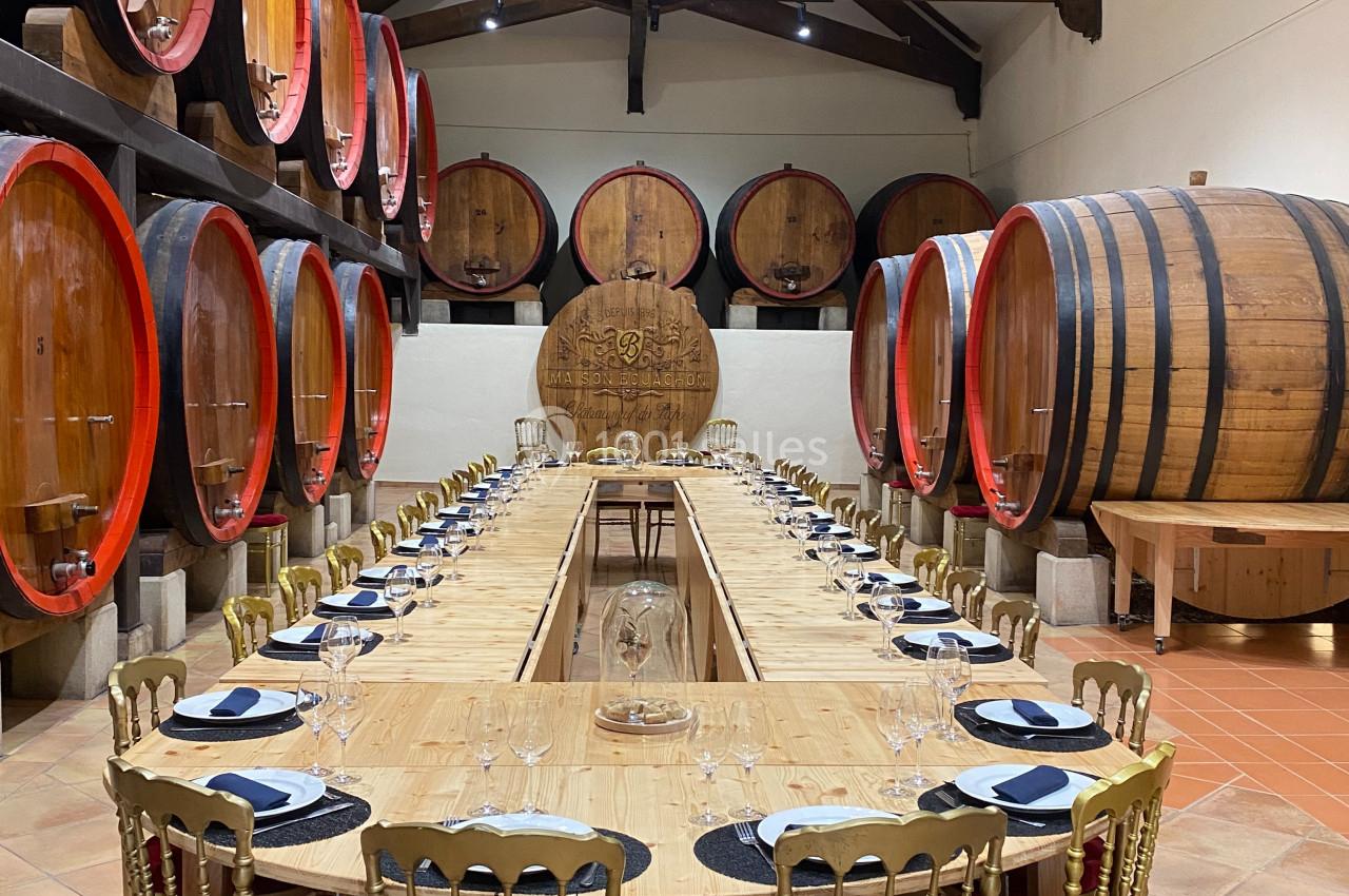 Salle de dégustation avec une longue table en bois entourée de chaises dorées, entourée de grandes barriques en bois.