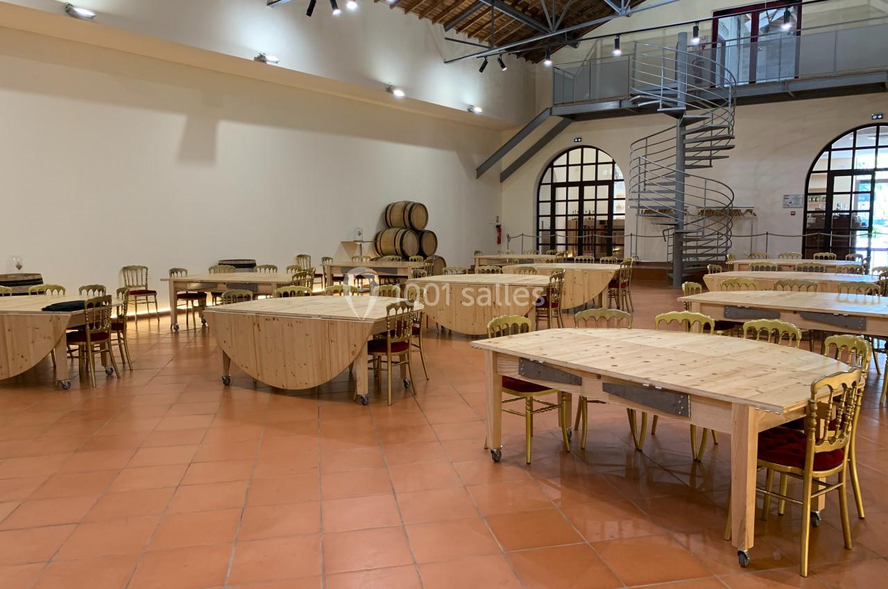 Salle spacieuse avec tables en bois pliantes, chaises dorées, tonneaux en arrière-plan et escalier en colimaçon.