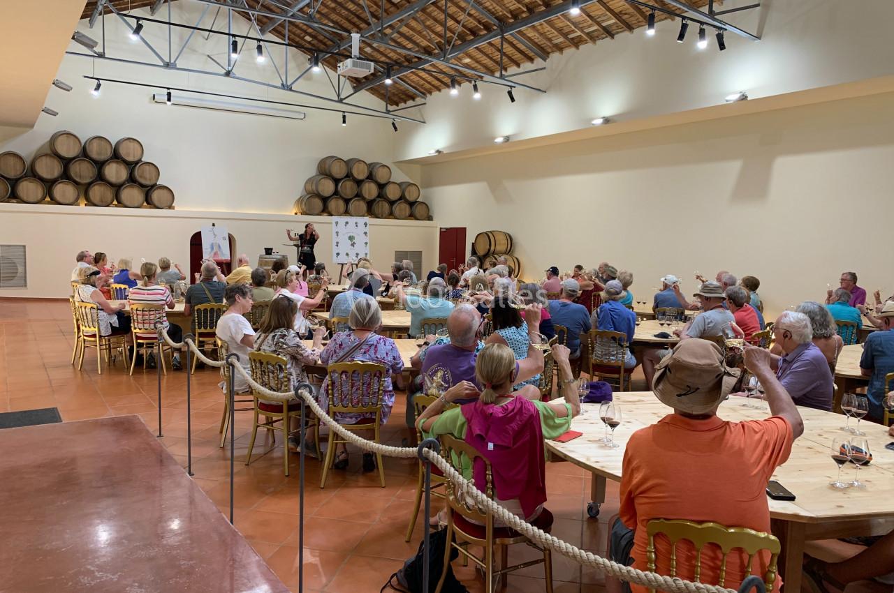 Groupe de personnes assises autour de tables dans une salle avec des tonneaux en arrière-plan, écoutant un intervenant.