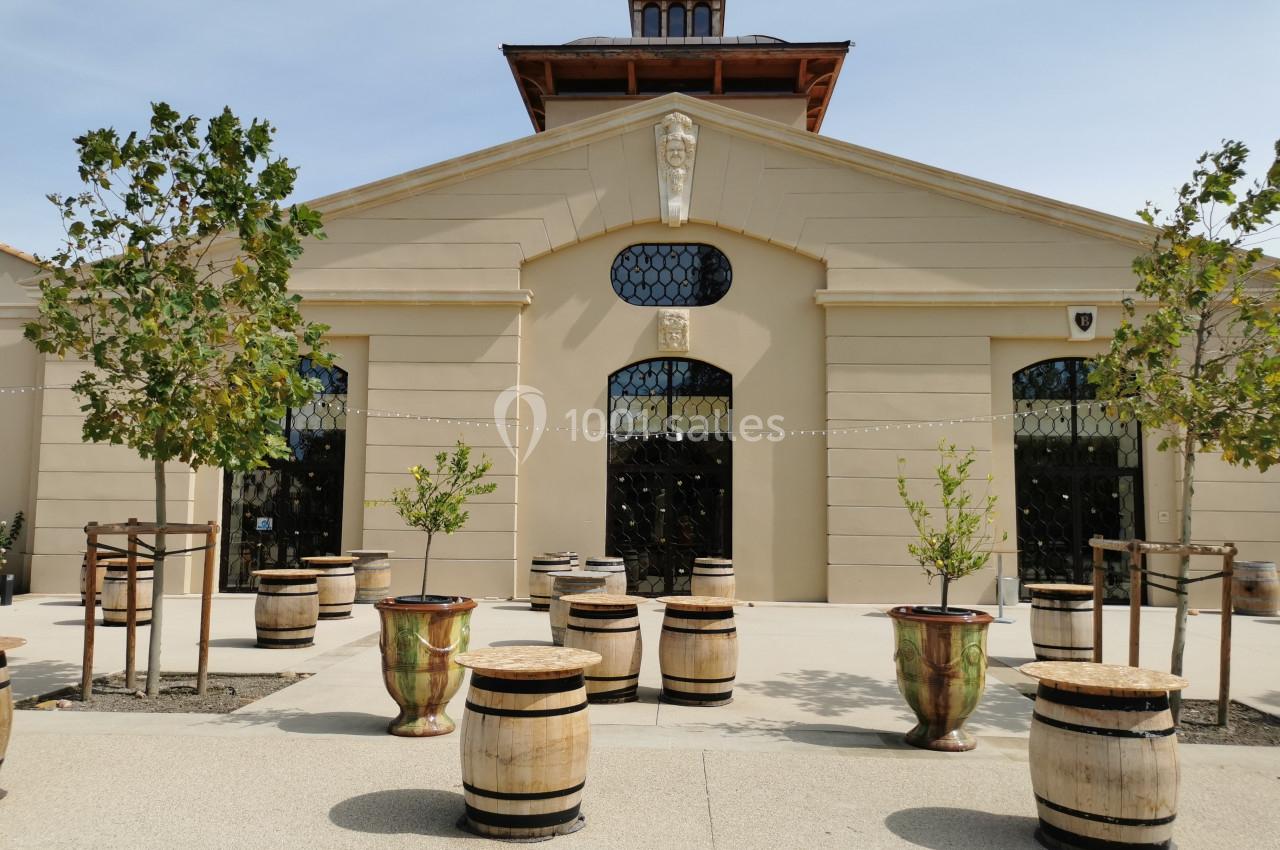 Façade d'un bâtiment beige avec des tonneaux en bois et des arbres plantés sur une esplanade pavée.