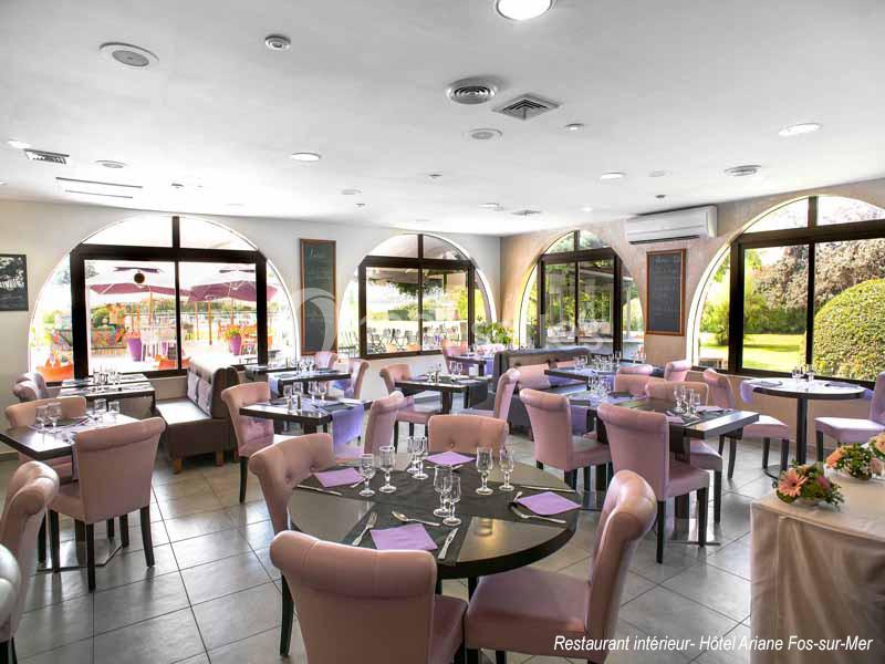 Salle de restaurant lumineuse avec tables dressées, chaises roses et grandes baies vitrées donnant sur une terrasse.