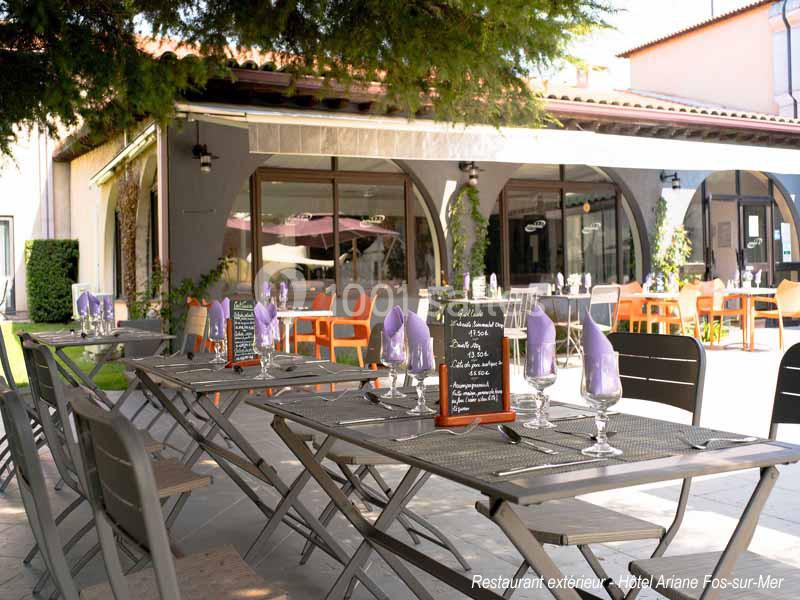 Terrasse d'un restaurant avec tables dressées, menus sur ardoises et vue sur une façade vitrée entourée de verdure.