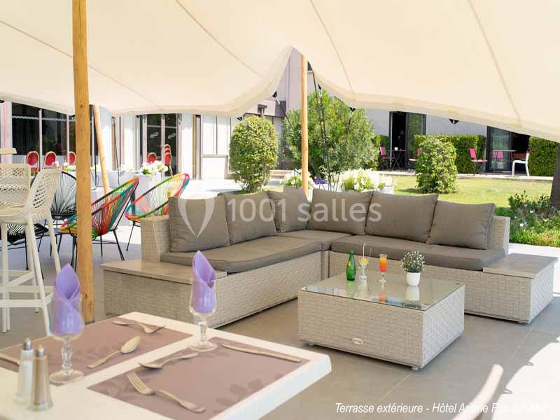 Salon de jardin avec canapé et table basse sur une terrasse ombragée, vue sur un jardin et des chaises colorées.