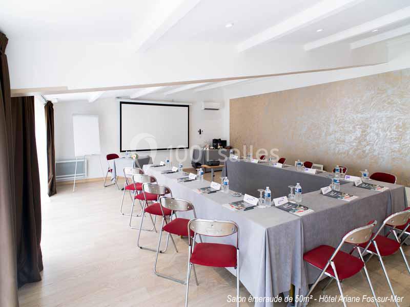 Salle de réunion lumineuse avec tables disposées en U, chaises rouges, écran de projection et paperboard.