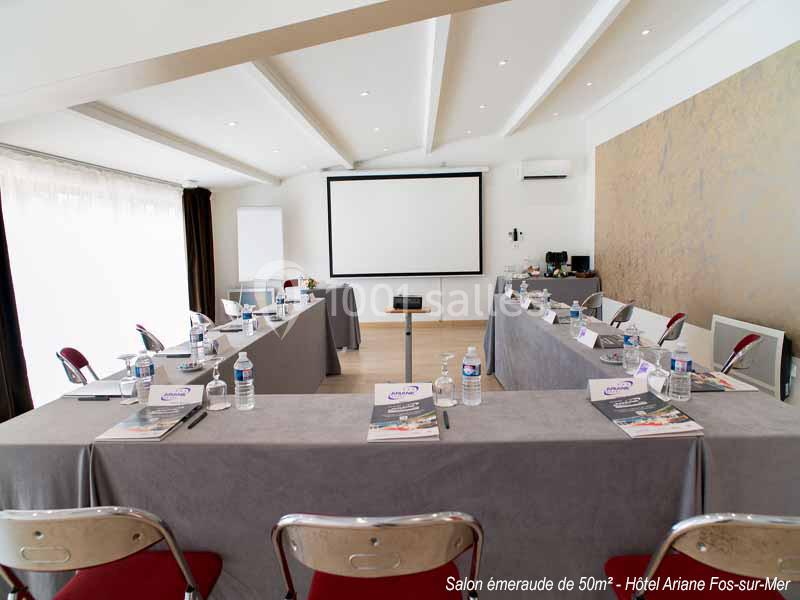 Salle de réunion lumineuse avec tables disposées en U, écran de projection, chaises et matériel de conférence.