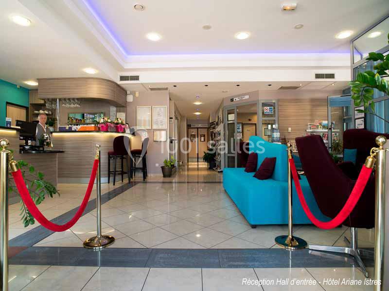 Hall d'entrée d'un hôtel avec comptoir de réception, coin salon bleu, plantes et cordons rouges délimitant l'espace.