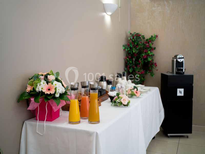 Table avec jus de fruits, café, tasses et décorations florales dans un espace intérieur lumineux.