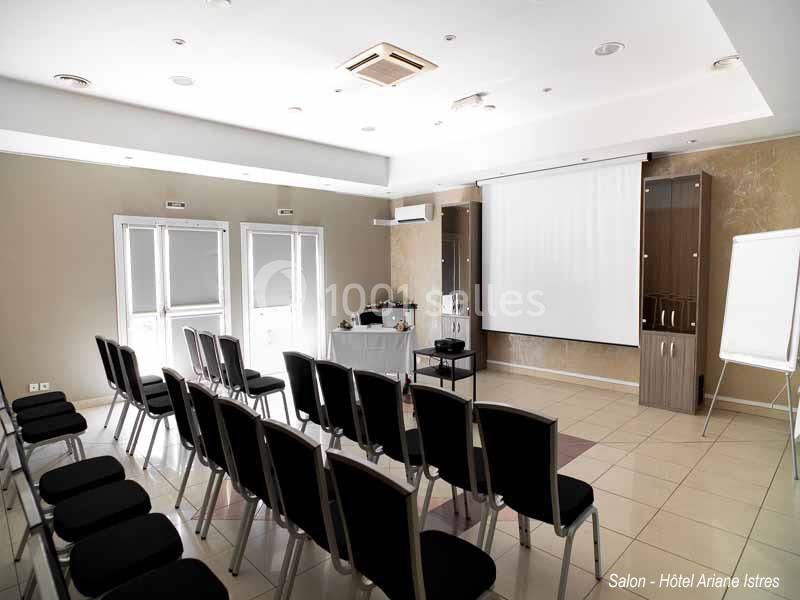 Salle de conférence lumineuse avec rangées de chaises, écran de projection, paperboard et équipement audiovisuel.
