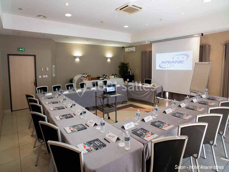 Salle de réunion équipée avec tables disposées en U, chaises, vidéoprojecteur, tableau blanc et documents.