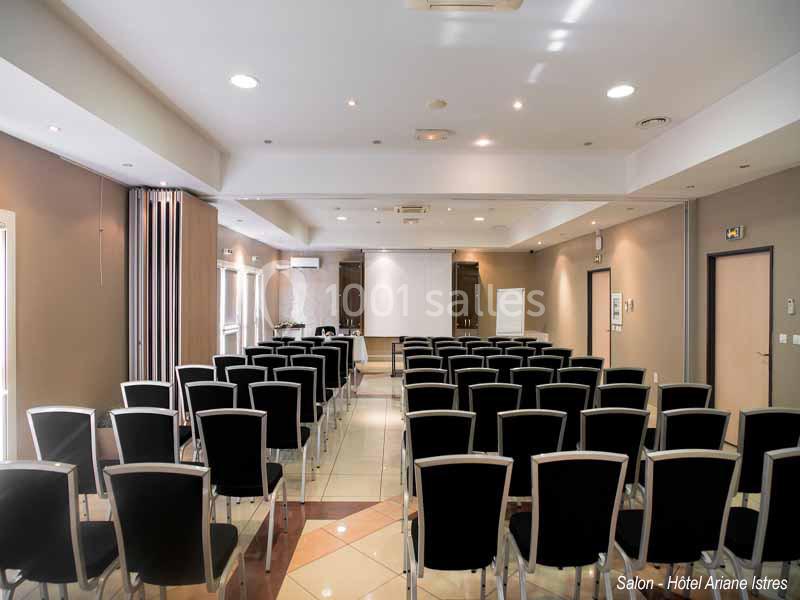 Salle de conférence lumineuse avec des rangées de chaises noires, un écran de projection et un tableau blanc.