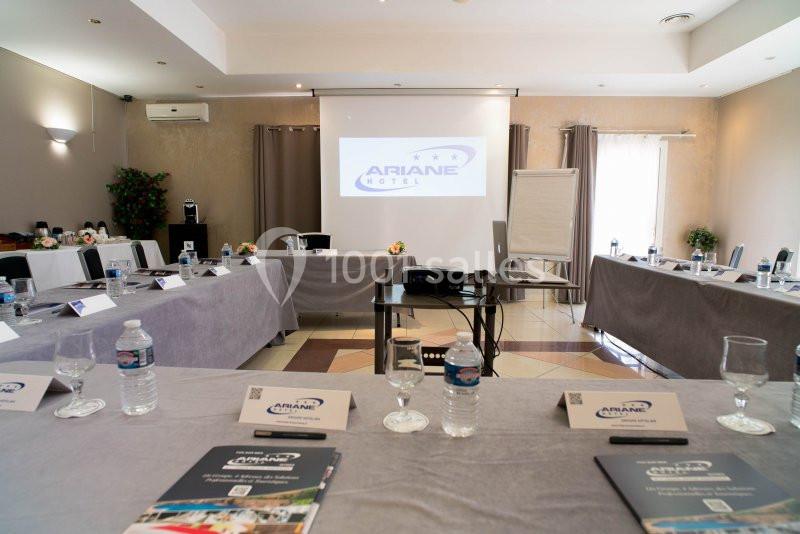Salle de réunion équipée avec tables disposées en U, vidéoprojecteur, écran, et fournitures de bureau.