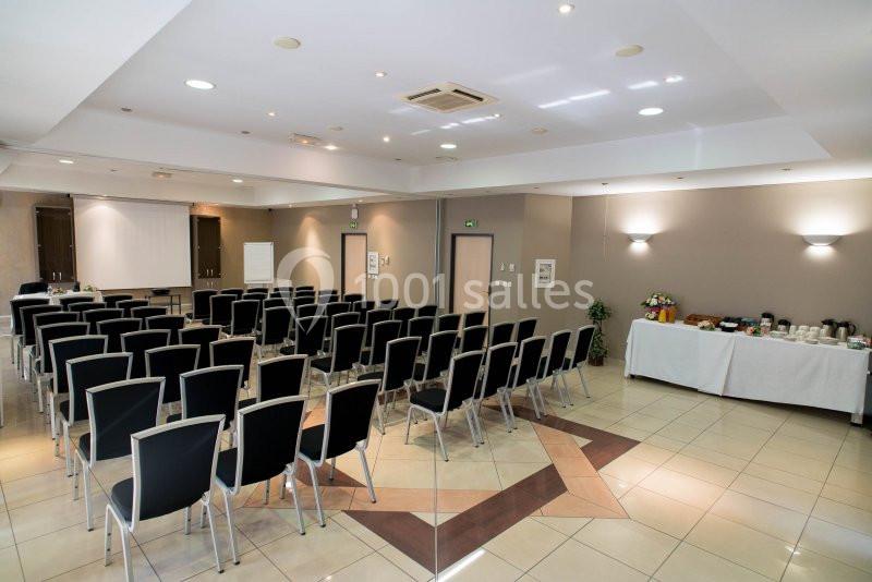 Salle de conférence avec rangées de chaises noires, écran de projection, paperboard et table de collation sur le côté.