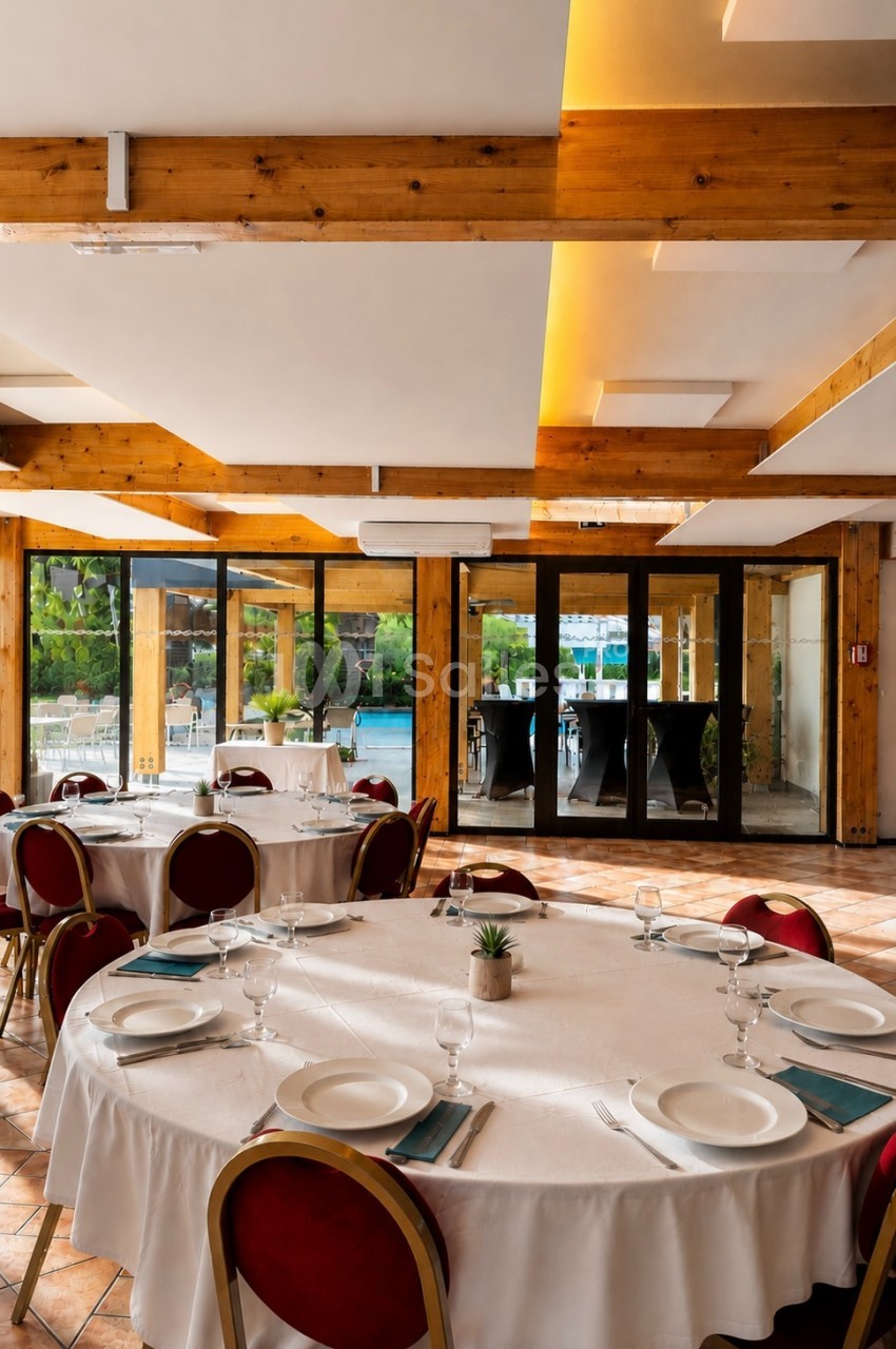 Salle de réception lumineuse avec tables rondes dressées, nappes blanches et vue sur une terrasse extérieure.
