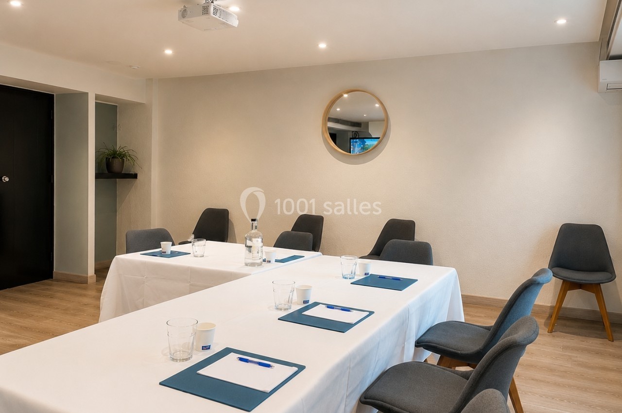 Salle de réunion lumineuse avec tables blanches, chaises grises, blocs-notes et un miroir rond au mur.