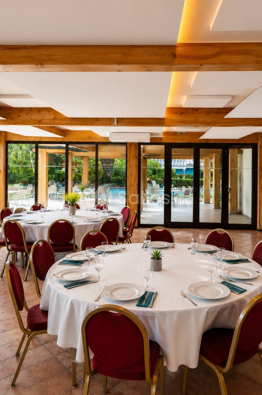 Salle de réception lumineuse avec tables rondes dressées, chaises rouges et vue sur un espace extérieur verdoyant.
