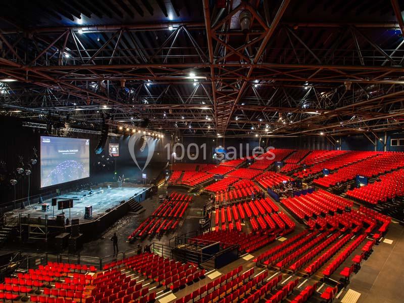 Salle de spectacle avec des rangées de sièges rouges, une scène éclairée et une structure métallique au plafond.