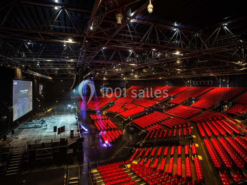 Salle de spectacle avec gradins rouges vides, scène éclairée et structure métallique apparente au plafond.