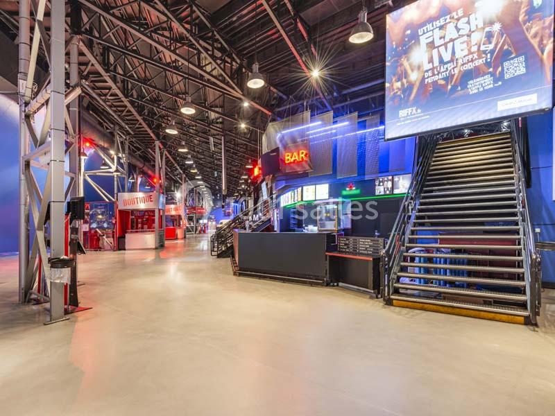 Intérieur d'une salle avec un bar, des escaliers et des enseignes lumineuses sous une structure métallique.