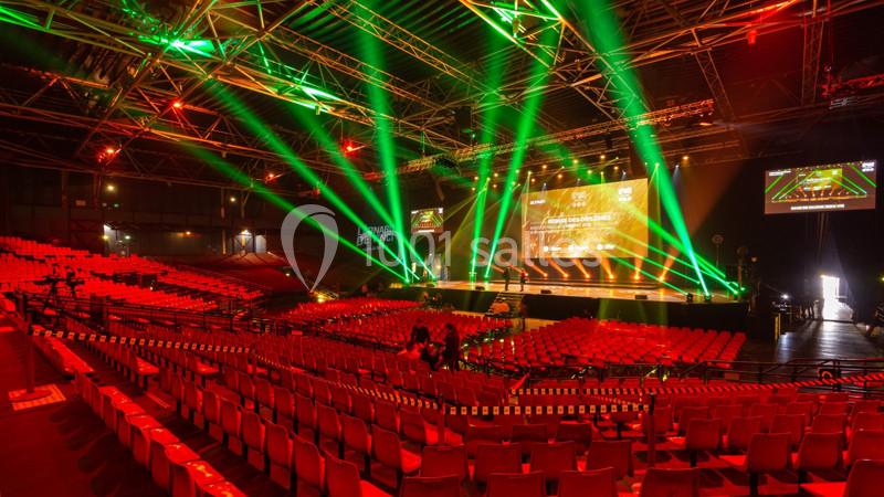 Salle de spectacle avec sièges rouges, scène éclairée par des projecteurs verts et rouges, et quelques personnes présentes.