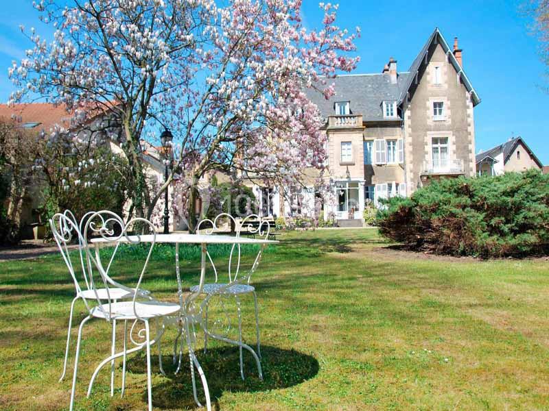 Table et chaises blanches en fer forgé sur une pelouse, devant une maison ancienne entourée d'arbres en fleurs.