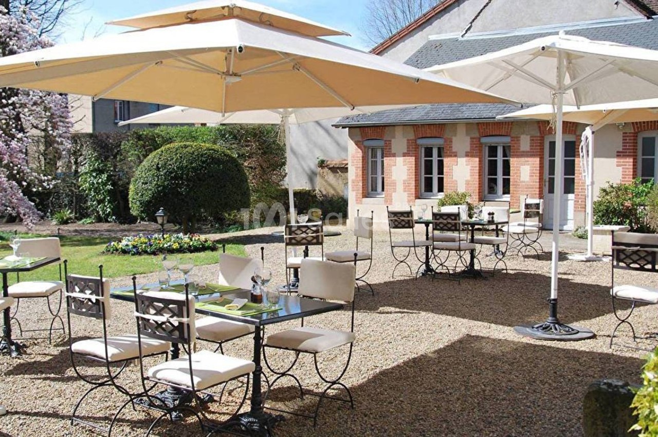 Terrasse extérieure avec tables et chaises en métal, parasols beige, jardin fleuri et bâtiment en arrière-plan.