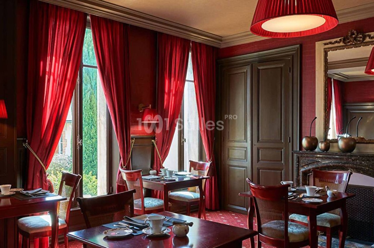 Salle à manger élégante avec rideaux rouges, tables dressées, cheminée et lumière naturelle entrant par de grandes fenêtres.