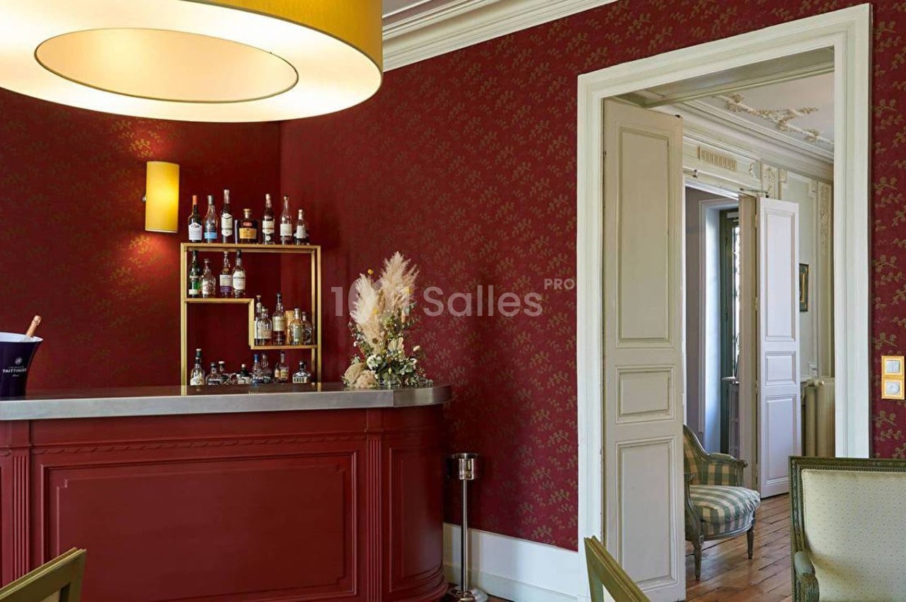 Un bar avec comptoir rouge, étagères de bouteilles, mur rouge orné, et vue sur une pièce adjacente lumineuse.