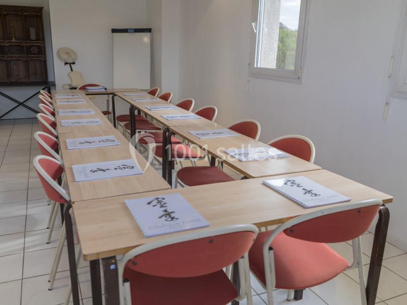 Salle de réunion lumineuse avec tables en U, chaises rouges et documents posés sur les tables.