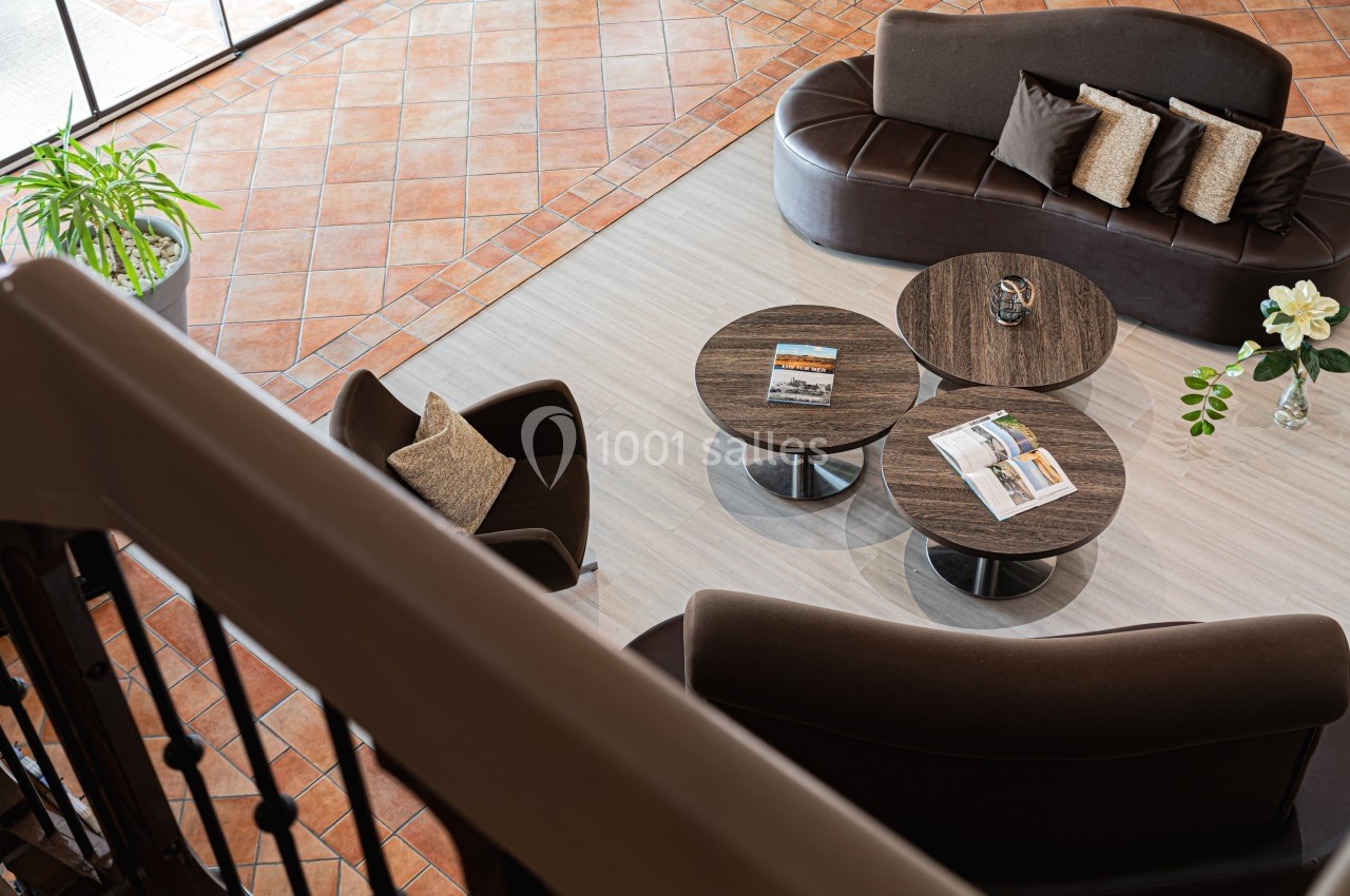 Vue d'un salon avec canapés marron, tables basses rondes, magazines et plantes sur un sol en carrelage et parquet clair.