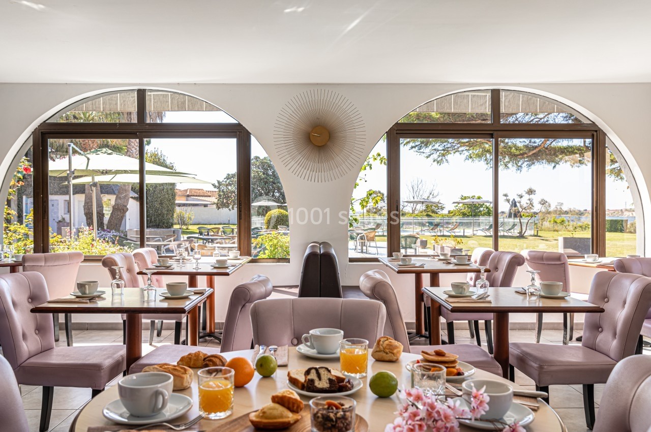 Salle lumineuse avec tables dressées pour le petit-déjeuner, vue sur un jardin et terrasse en arrière-plan.