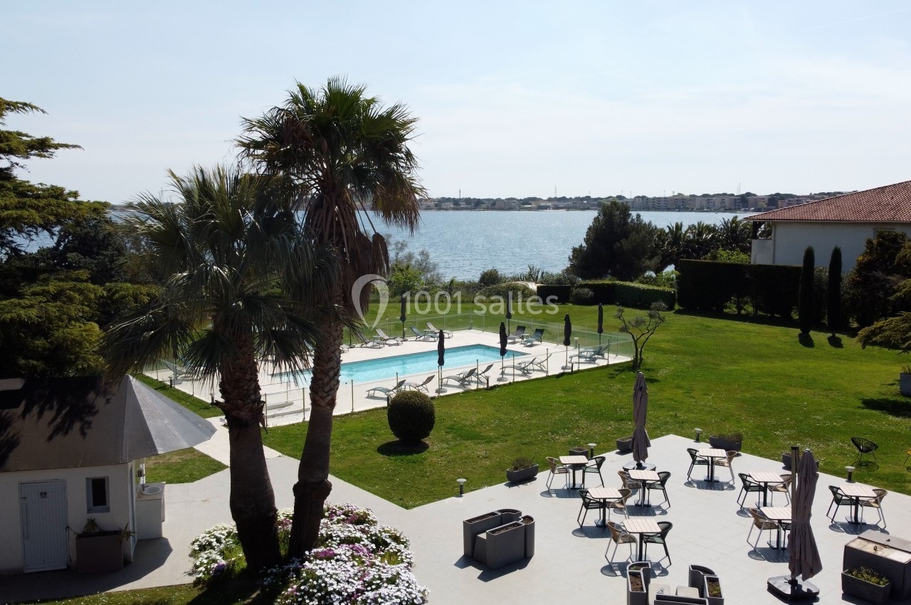 Vue d'une piscine extérieure entourée de transats et de verdure, avec un paysage côtier en arrière-plan.