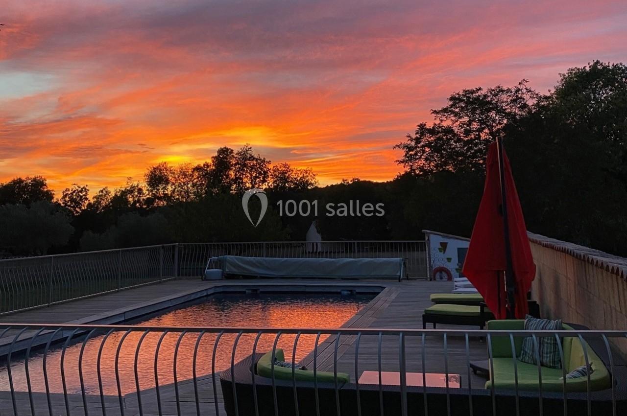 Piscine extérieure entourée de transats et parasols rouges, au coucher du soleil avec un ciel aux teintes orangées.