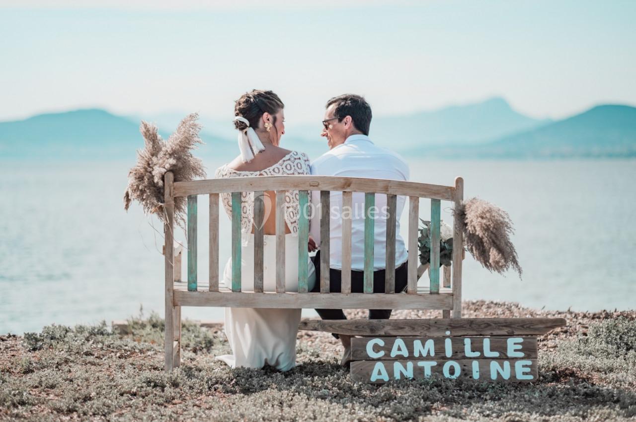 Un couple assis sur un banc en bois près d'un lac, entouré de pampas, avec une pancarte portant leurs prénoms.