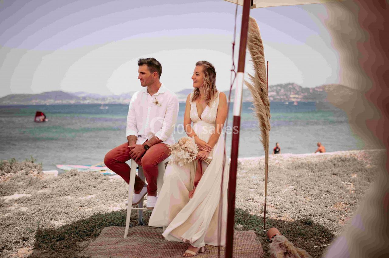 Un couple assis sous un parasol près de la mer, entouré d'herbe et d'éléments décoratifs naturels.