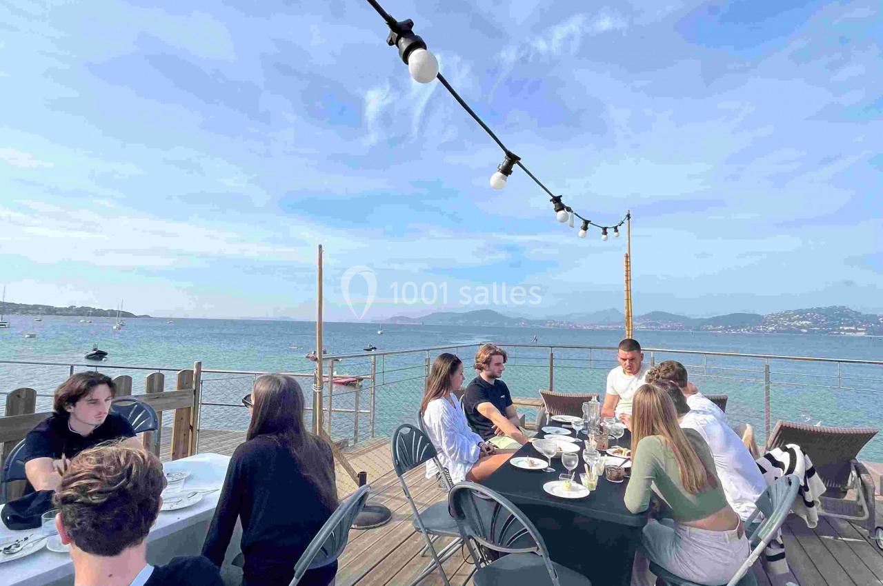 Des personnes assises autour de tables sur une terrasse en bois, avec vue sur la mer et un ciel dégagé.